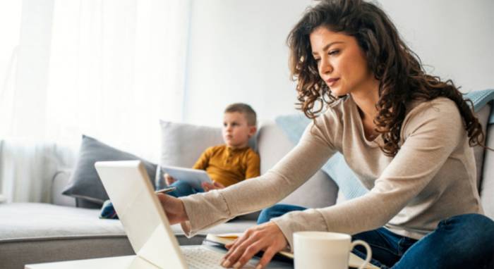 Femme travaillant sur ordinateur portable depuis son domicile, avec un enfant sur son canapé